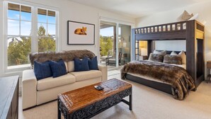 A modern bedroom with a bunk bed, a sofa, a coffee table, and a large window offering a view of trees and mountains. - Ski-In/Ski-Out via Pony Express Lift (Big Sky)