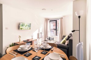 A dining area with a round table set for four, a sofa, a TV mounted on the wall, and a window with curtains.