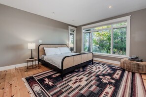 Une chambre à coucher comprenant un lit, une table de chevet, une lampe, une fenêtre donnant sur la forêt et un tapis à motifs.
