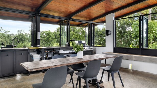 An open-plan kitchen with a wooden table and chairs, large windows, and a view of greenery.