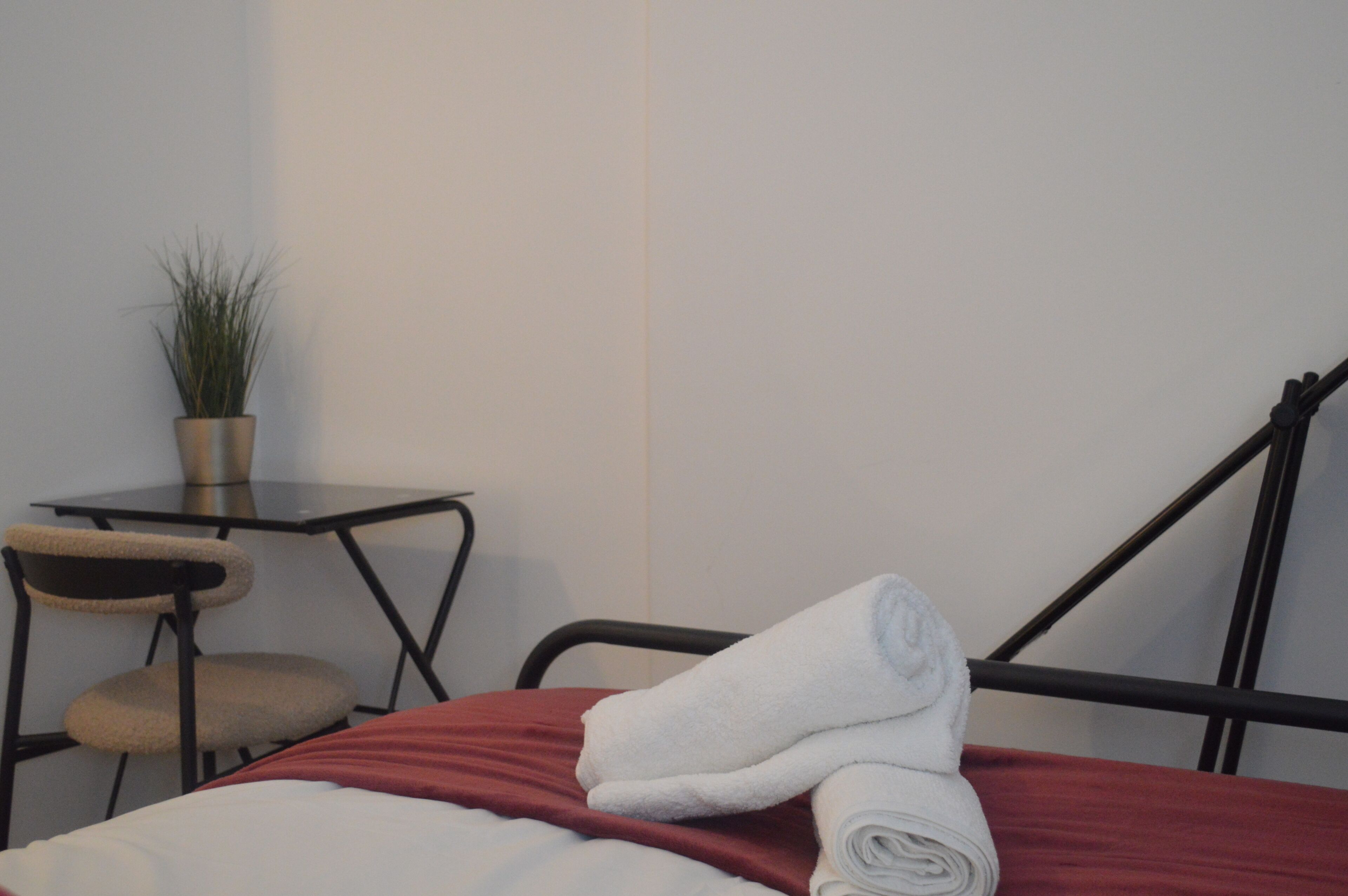 A neatly made bed with a red blanket and a rolled white towel, a small table with a potted plant, and a chair.