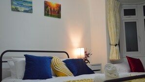 Ein ordentlich bezogenes Bett mit blauen und gelben Kissen, einer weißen Decke und einem roten Kissen. An der Wand hängt ein Gemälde, das einen Sonnenuntergang zeigt, ein Nachtlicht steht auf dem Nachttisch und ein Fenster mit Vorhängen ist zu sehen.