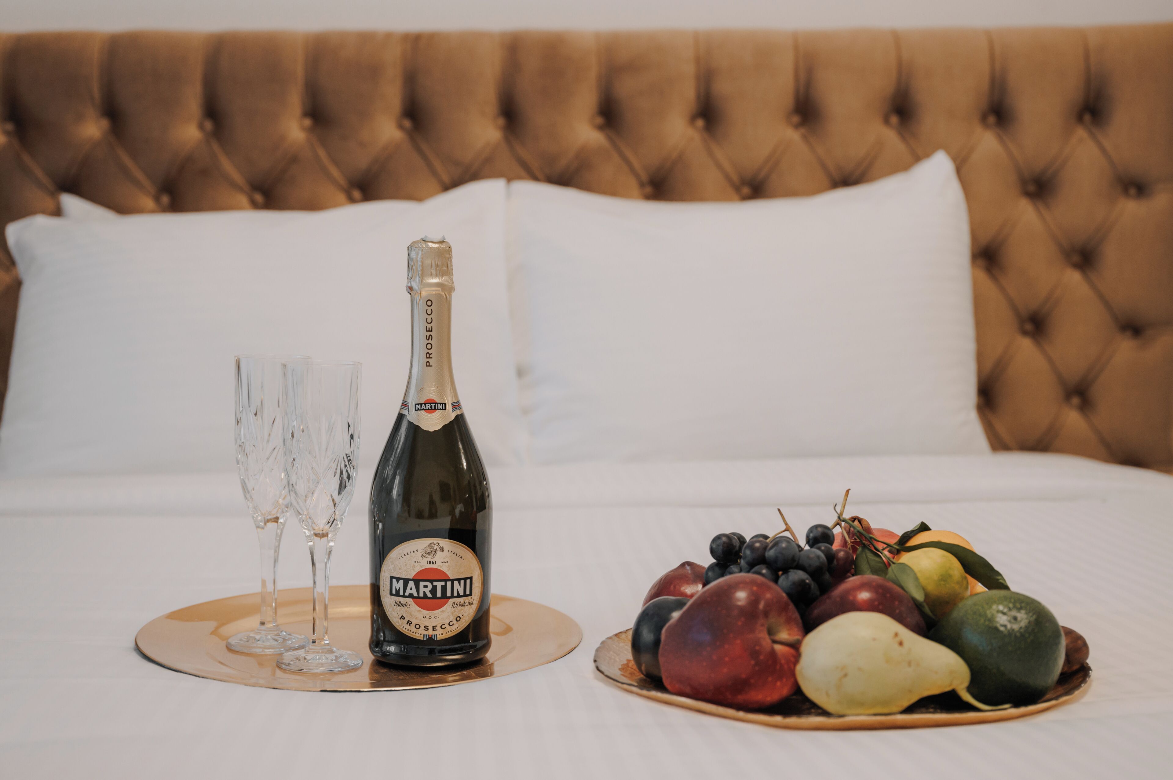 A bed with a tufted headboard, a bottle of Martini wine, two glasses, and a fruit platter on a white bedspread.