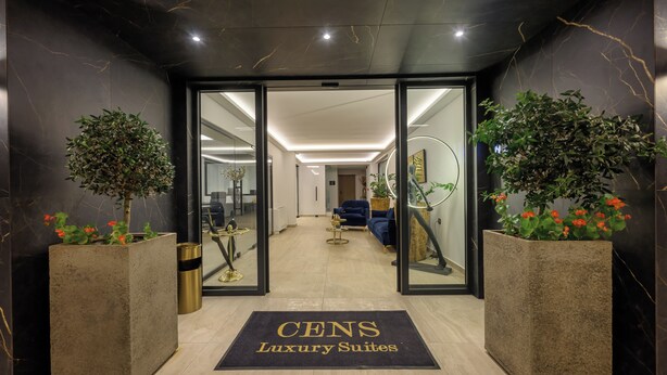 Entrada de un hotel con felpudo que dice "CENS Luxury Suites", un interior moderno con un sofá azul y una planta, y una pared de mármol.