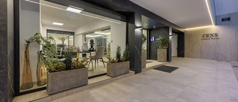 A modern hotel entrance with glass doors, indoor plants, and a sign for 'CENS Luxury Suites'.