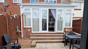 A patio area with a brick wall, a white-framed window, and a small step leading to a door. There are two outdoor chairs and a table. - 3 Bedroom - 1 Bathroom - Garden - Pet Friendly (Wrexham)