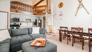Ein modernes Wohnzimmer mit einer grauen Eckcouch, einem Holzespeisetisch und Stühlen. Im Hintergrund ist eine Küche mit weißen Schränken und einer Mikrowelle erkennbar.