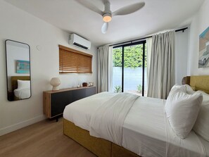 A bedroom with a bed, bedside table, mirror, air conditioning unit, window with curtains, and a ceiling fan.