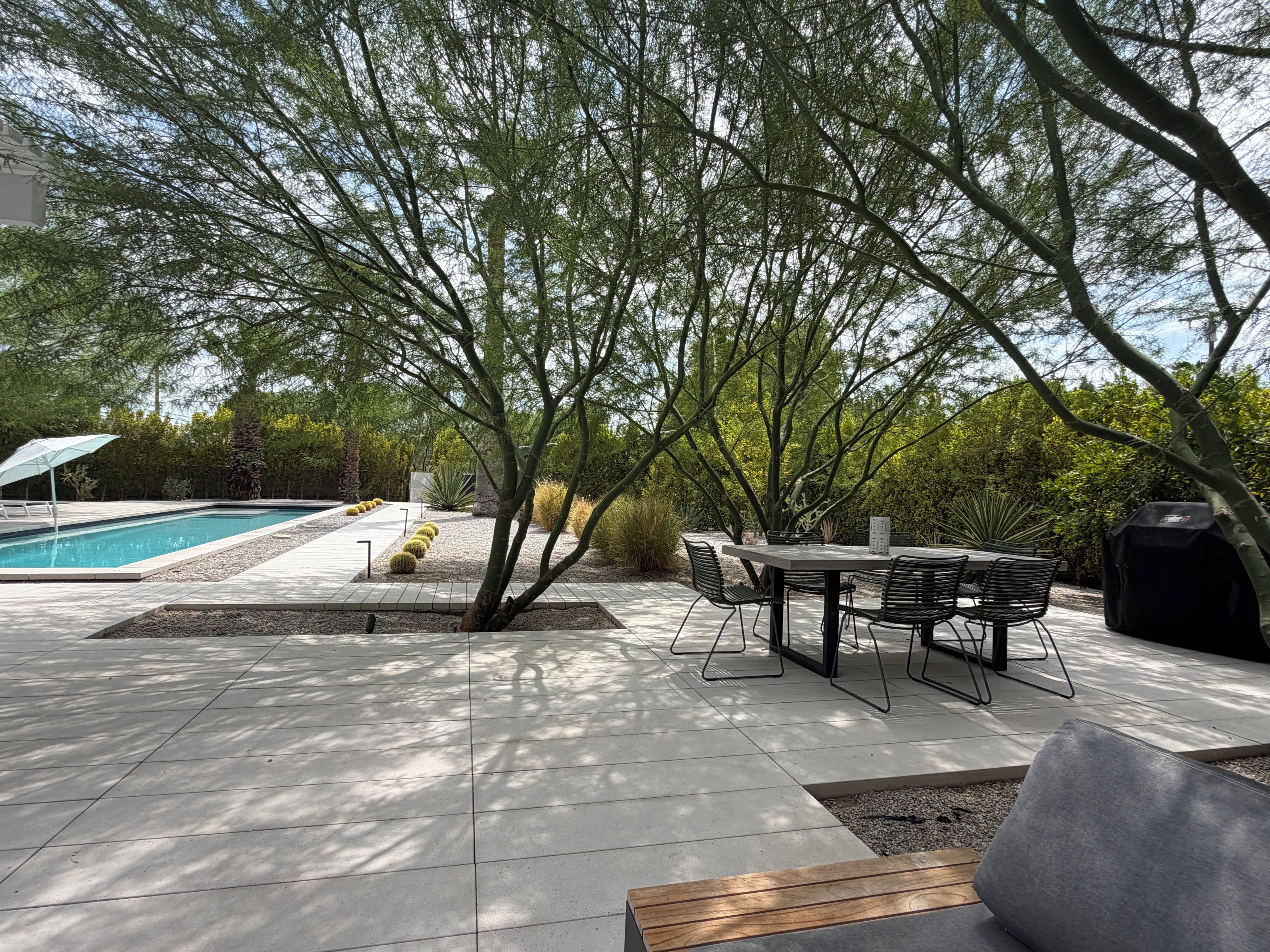 A poolside area with a table and chairs, surrounded by trees and a barbecue grill.