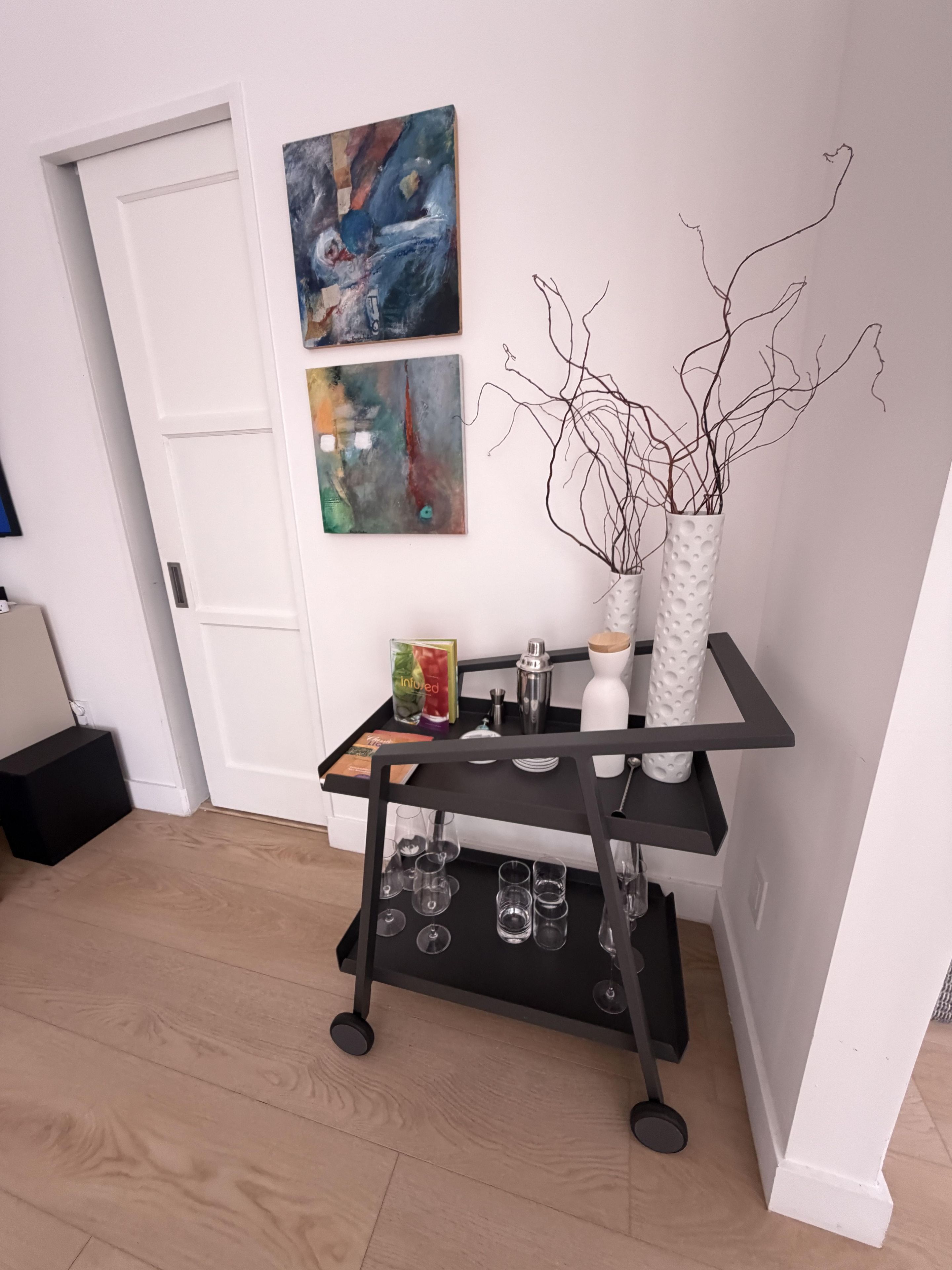 Een kamer met een zwarte metalen wagen waarop glazen en een witte vaas met takken staan. Aan de muur hangen abstracte schilderijen.