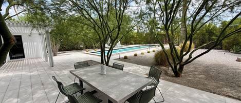 Une terrasse extérieure avec une table et des chaises, entourée d’arbres et bordée d’une piscine.