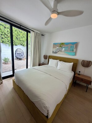 A bedroom with a bed, a ceiling fan, a nightstand, a wall-mounted lamp, and a window with curtains. - The Knot - The Ultimate Palm Springs Bungalow (Palm Springs)