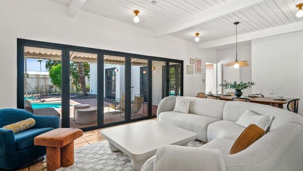 Una sala moderna con un sofá seccional grande, una mesa de centro y un sillón azul. Hay un comedor con una mesa y sillas, y vistas a una piscina y un área de estar al aire libre.