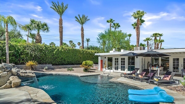 A swimming pool with lounge chairs and umbrellas, surrounded by palm trees and a building with large windows.