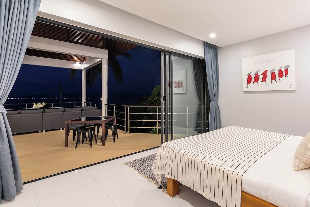 A modern bedroom with a large bed, a balcony with a dining area, and a city view at night.