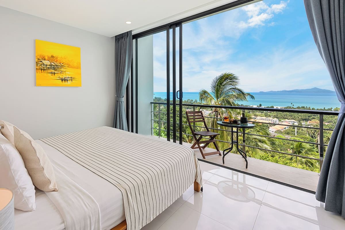 A bedroom with a large bed, a nightstand, and a view of a balcony with a table and chair.