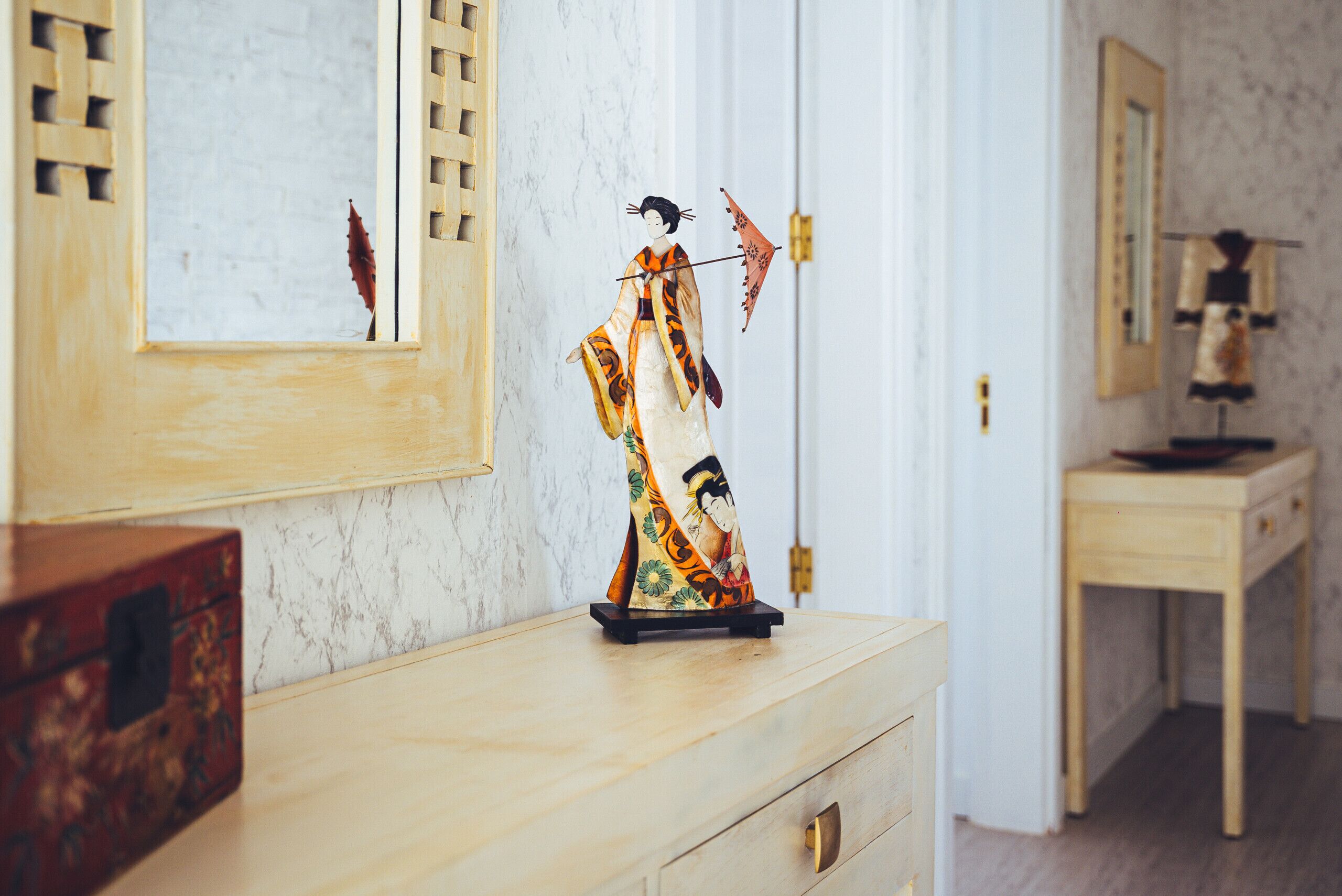 Eine traditionelle japanische Puppe auf einer Kommode, ein Spiegel an der Wand und ein Holzschrank.