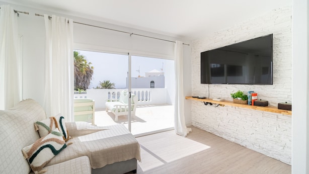 A modern living room with a sofa, a flat-screen TV mounted on a white brick wall, and a sliding glass door leading to an outdoor balcony with a view of buildings and palm trees.
