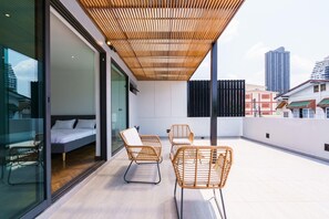 A modern balcony with a bed, two wicker chairs, and a view of the cityscape.