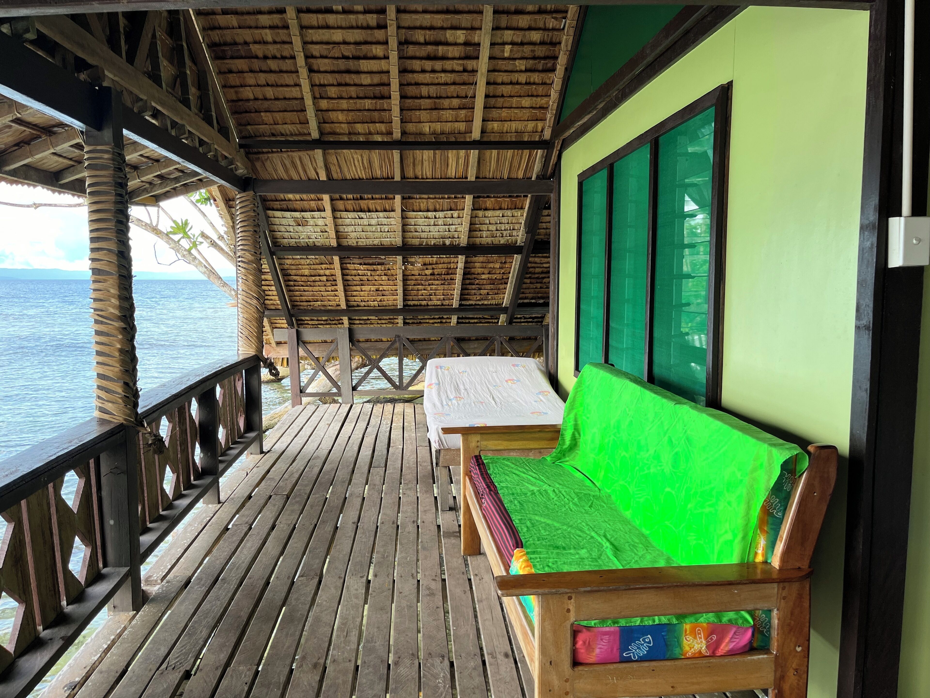 A wooden deck with a thatched roof, a green wall, and a bench with colorful cushions.