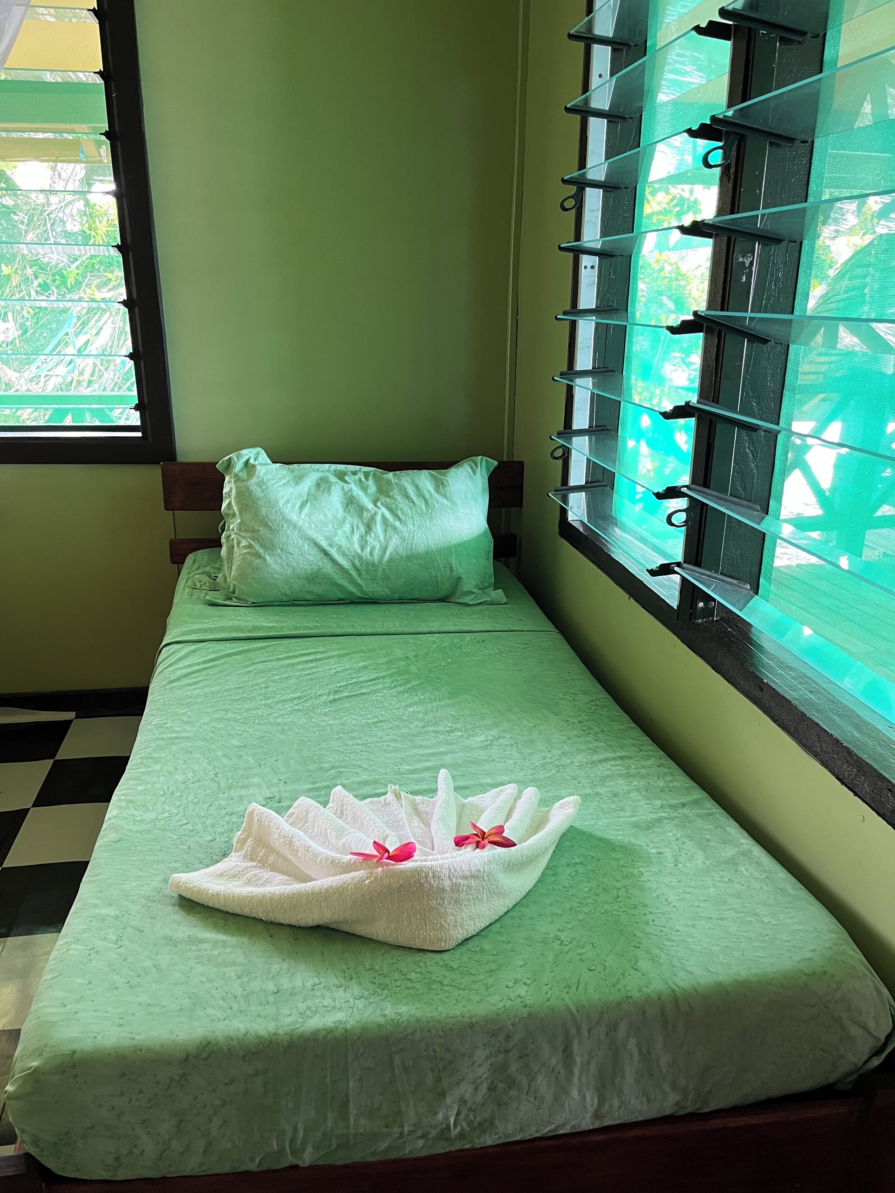 Una cama individual con una colcha verde y una almohada blanca, una toalla blanca pequeña con flores rosas, y una ventana con mosquitera verde.