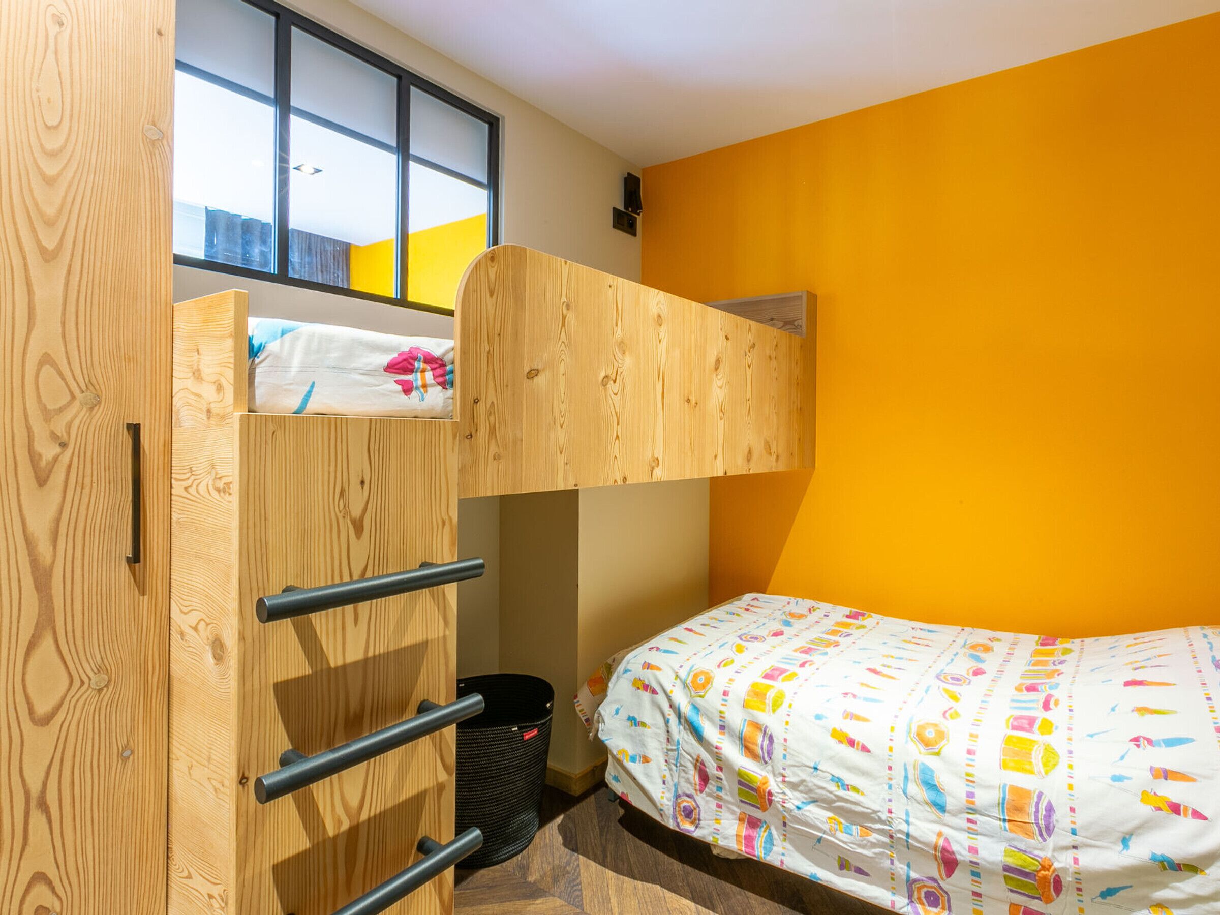 A bunk bed room with a window, a wooden wardrobe, and a colorful bedspread.