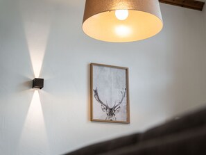 Uma obra de arte com cabeça de veado emoldurada em uma parede branca, um aparelho de luz fixado na parede e uma viga de madeira acima.