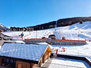 En træbygning med et skråt tag, sandsynligvis en hytte eller restaurant, ligger ved siden af en sneklædt skibakke med stolelifter og en klar blå himmel.