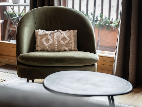 Un fauteuil vert agrémenté d’un coussin à motifs, une petite table ronde et une vue sur un balcon orné de plantes.