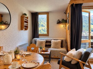 A cozy room with a wooden table set for dining, a sofa with a view of the mountains, and a window with curtains. - Renovated ski-in studio with balcony and Wi-Fi (Méribel)