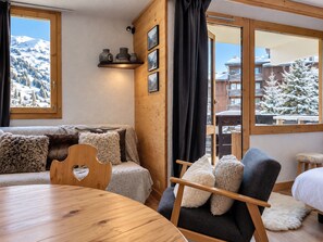 A cozy room with a wooden ceiling, a sofa with fluffy cushions, a wooden chair, a round wooden table, and a window with a view of snow-covered trees and buildings. - Renovated ski-in studio with balcony and Wi-Fi (Méribel)