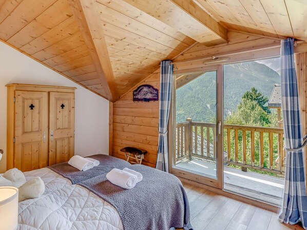 A wooden cabin room with a bed, a small table, and a large window offering a mountain view.