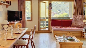 Ein gemütliches Zimmer mit Holztisch, gedecktem Esstisch, einer Couch mit rotem Kissen, einem Fernseher und einem großen Fenster mit Blick auf eine Berglandschaft.