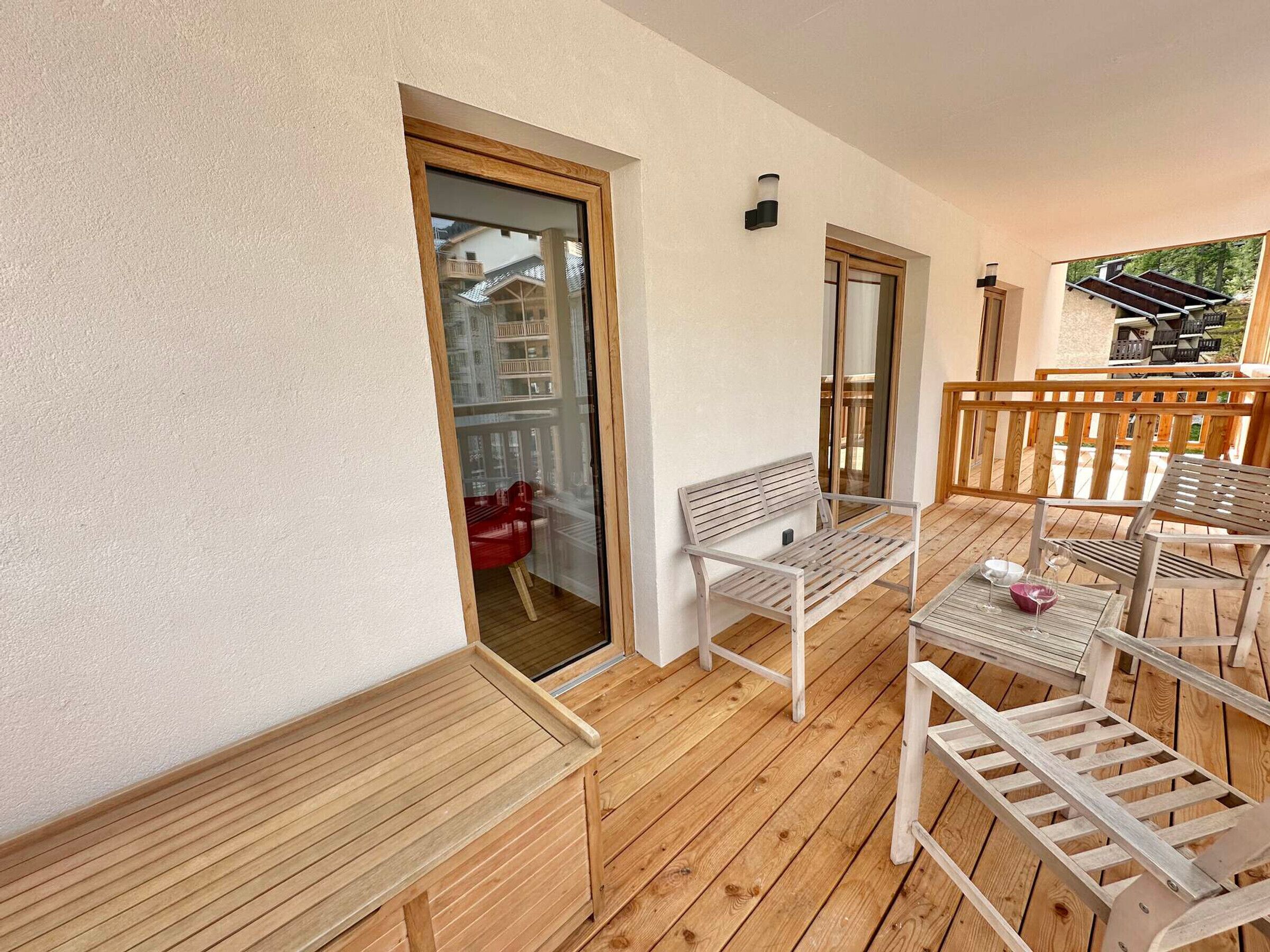 Eine Holzterrasse mit Bank und Tisch, eine Glastür zum Verschieben und Blick auf ein Gebäude im Hintergrund.