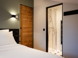 A hotel room with a bed, a wooden door, a glass shower enclosure, and a wall-mounted light fixture.