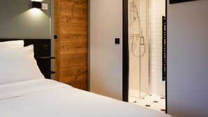 A hotel room with a bed, a wooden door, a glass shower enclosure, and a wall-mounted light fixture. - 4★, 5 pers, village center, 125 m slopes, 3 bedrooms, south balcony, parking (La Clusaz)