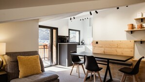 A modern living room with a sofa, a dining table with chairs, a kitchen area, and a balcony with a view. - 4★, 5 pers, village center, 125 m slopes, 3 bedrooms, south balcony, parking (La Clusaz)
