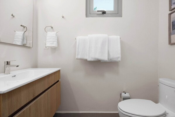 Un baño con un lavamanos blanco, un armario de madera, dos toallas colgadas en ganchos, un inodoro y una ventana pequeña.