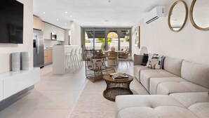 A modern living room with a sofa, coffee table, and wicker chairs. A kitchen with a refrigerator and microwave is visible in the background.