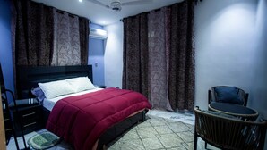 A bedroom with a bed, a chair, a small table, a ceiling fan, and an air conditioning unit.