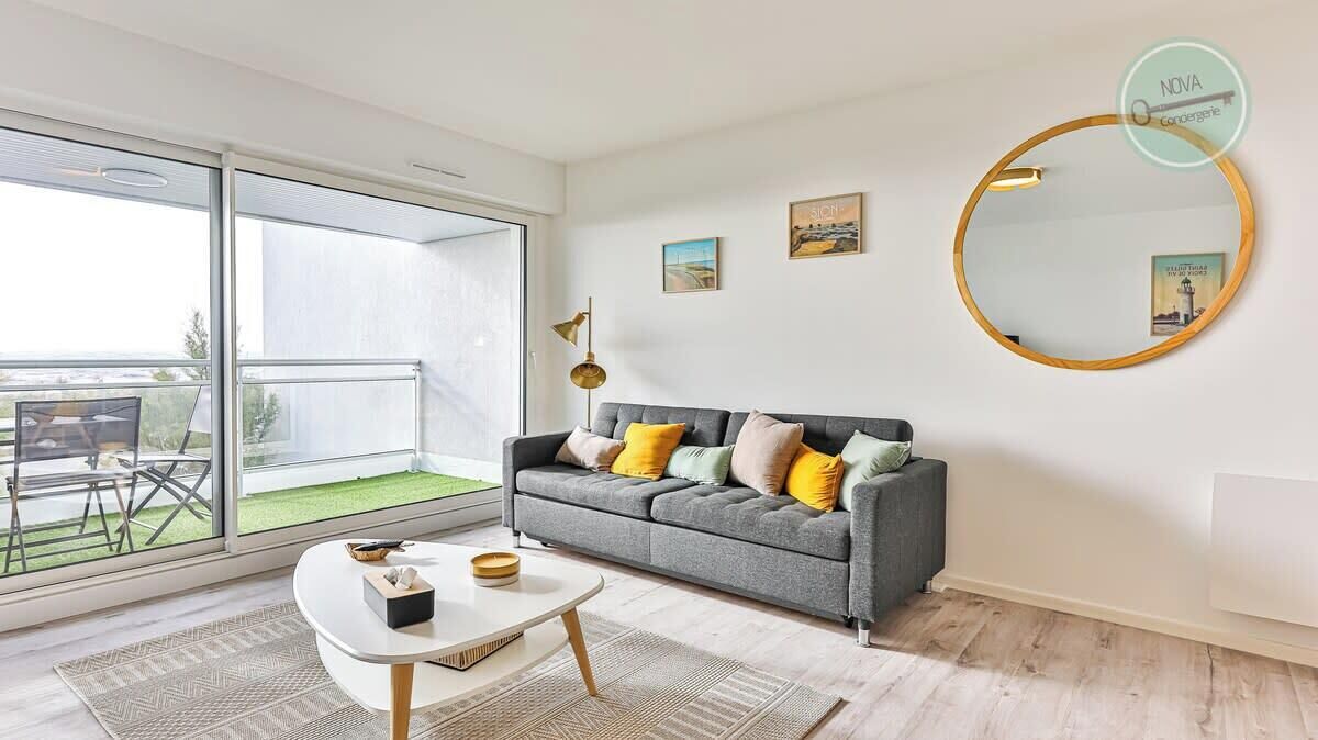 A modern living room with a grey sofa, yellow pillows, a round mirror, and a view of a balcony with a green lawn.