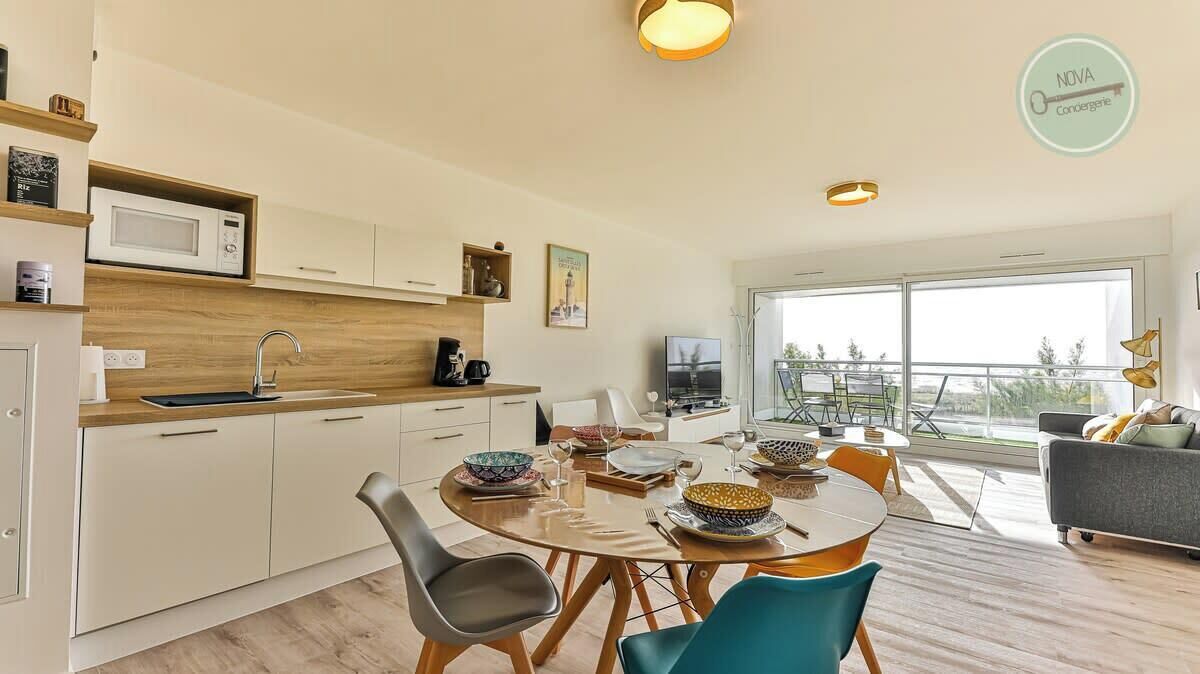 A modern kitchen with white cabinets, a microwave, and a coffee maker. There is a dining area with a round table and chairs, and a sofa in the living room.