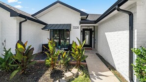 A white brick building with a black roof, a number '7006' on the wall, and a small garden area with plants and a bench. - Cozy 3-bedroom house in welcoming Arlington with WiFi, AC (Arlington)