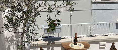 A terrace with a potted olive tree, wooden chairs, a table, and a bottle of wine.
