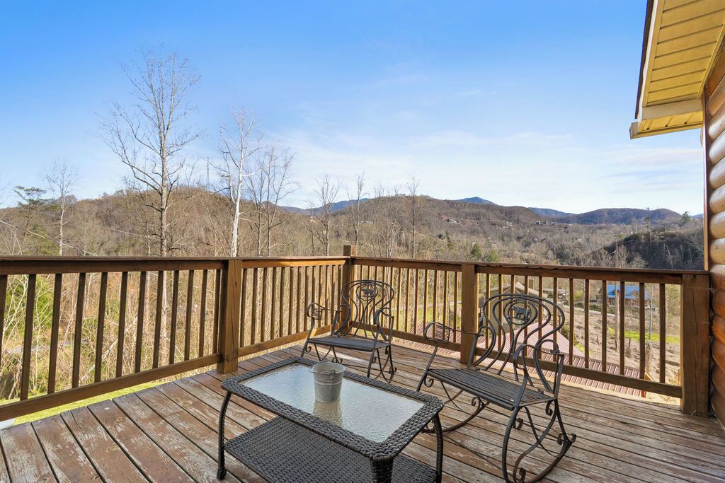 A wooden deck with a glass table and metal chairs, overlooking a hilly landscape.