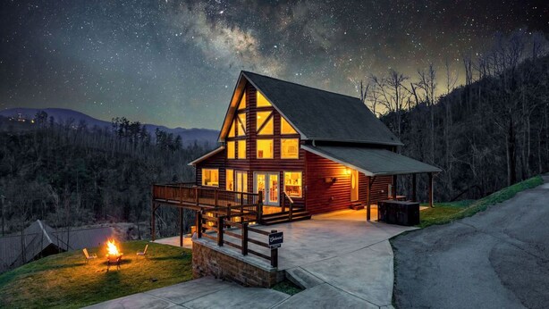 A log cabin with a porch, a fire pit, and a view of the night sky.