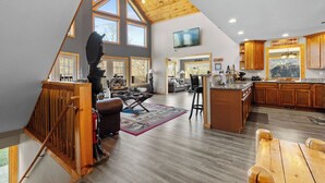 A spacious loft-style living area with a high ceiling, large windows, a wooden staircase, a dining area, and a kitchen with wooden cabinets.