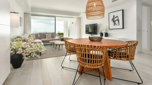 A modern living room with a round wooden dining table, wicker chairs, a large window, a sofa, and a TV. - Luxury 2-bedroom sea View Apartment in Casares (Casares)