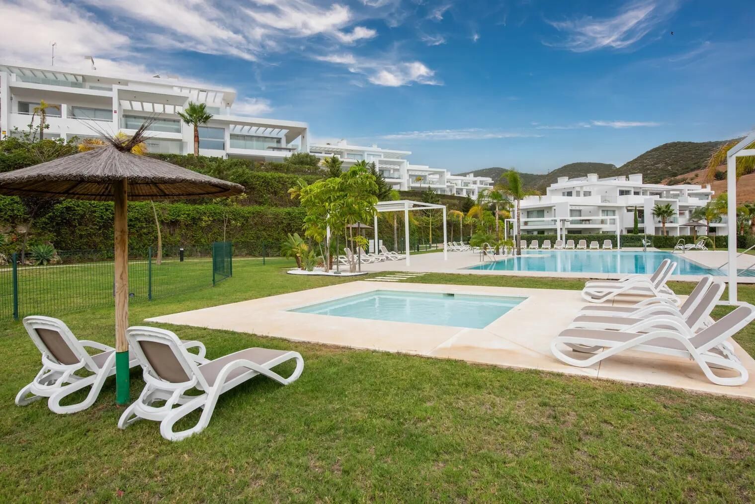 A pool area with lounge chairs, a thatched umbrella, and a view of modern white buildings.