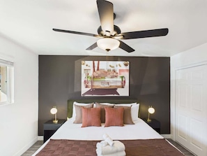 A bedroom with a bed, a ceiling fan, a wall-mounted artwork, bedside lamps, and a door.